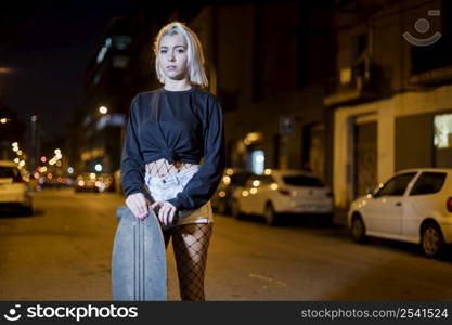 Young pretty woman looking camera in shorts while standing on the street holding a skateboard at night in the city