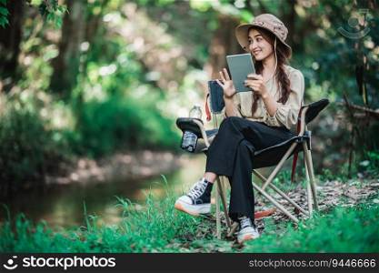 Young pretty woman sitting in chair and use tablet video call while camping in nature park, copy space