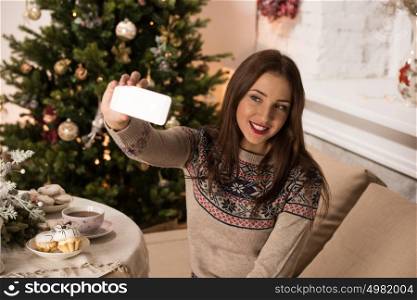Young pretty woman taking selfie at home while sitting on sofa near Christmas tree