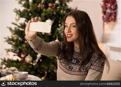 Young pretty woman taking selfie at home while sitting on sofa near Christmas tree