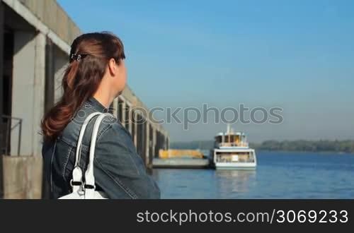 young pretty woman watching on river from platform near shore, boat at background in defocus