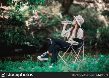 Young pretty woman wearing c&ing hat and sitting on chair, She drink water in bottle near stream while c&ing in nature forest, copy space