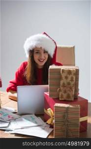 Young pretty woman wearing Santa Claus hat wrapping Christmas gift at her desk and responding to children messages and wishes by email