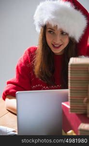 Young pretty woman wearing Santa Claus hat wrapping Christmas gift at her desk and responding to children messages and wishes by email