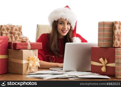 Young pretty woman wearing Santa Claus hat wrapping Christmas gift at her desk and responding to children messages and wishes by email