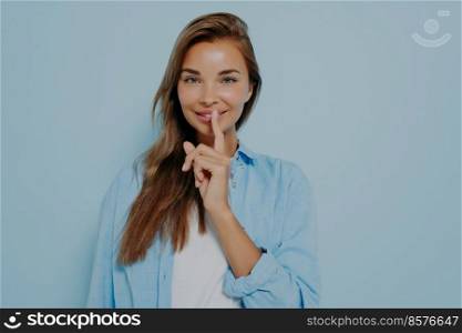 Young pretty woman wide eyed with smile asking for silence or secrecy with finger on lips with hush hand gesture isolated on blue background. Positive emotions and facial expression, body language. Pretty female placing fingers on lips making sign symbol