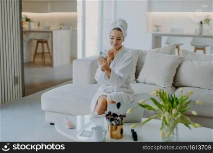 Young pretty young woman looks at modern smartphone camera takes selfie has well cared healthy skin applies cream wears bathrobe and towel poses on sofa does cosmetic procedures for complexion