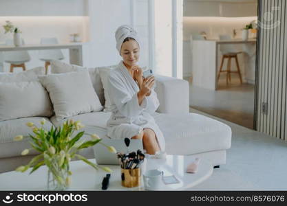 Young pretty young woman looks at modern smartphone camera takes selfie has well cared healthy skin applies cream wears bathrobe and towel poses on sofa does cosmetic procedures for complexion