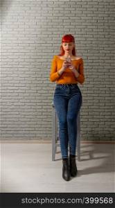 Young red-haired girl is reading something thoughtfully on her smartphone against a gray brick wall. girl watching the phone