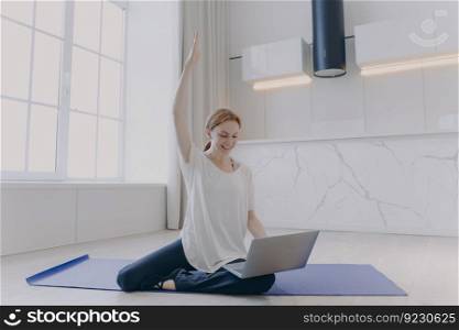 Young red haired woman in sportswear sitting on her knees on floor with laptop. Practicing yoga on mat. Online fitness tutorial. Concept of home classes and distance training on quarantine.. Young woman sitting on her knees on floor with laptop. Online fitness tutorial. Distance training.