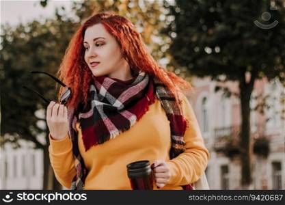 Young red-haired woman in yellow sweater and sunglasses holds thermo mug with coffee in her hands. Lady walks through city and park on sunny autumn day. Eco-friendly lifestyle.. Young red-haired woman in yellow sweater and sunglasses holds thermo mug with coffee in her hands. Lady walks through city and park on sunny autumn day.