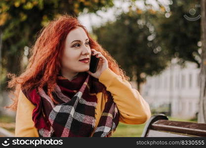 Young red-haired woman in yellow sweatshirt talking on the phone. Lady sits on park bench on fall day. Lifestyle.. Young red-haired woman in yellow sweatshirt talking on phone. Lady sits on park bench on fall day. Lifestyle.