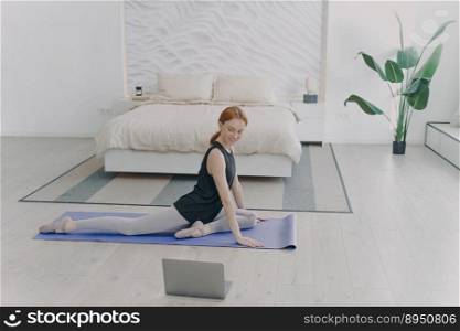 Young red haired woman practicing yoga by video lessons on laptop. Girl is doing workout and stretching in her bedroom. Concept of internet learning and home classes.. Red haired woman practicing yoga by video lessons in bedroom. Internet learning and home classes.
