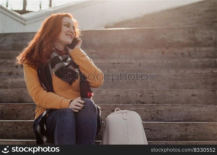 Young red-haired woman sitting on stairsand talking on phone. Lady in yellow sweater uses smartphone on sunny day. Lifestyle, copy space.. Young red-haired woman sitting on stairs and talking on phone. Lady in yellow sweater uses smartphone on sunny day. Lifestyle