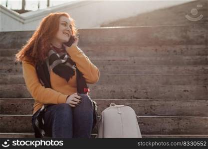 Young red-haired woman sitting on stairsand talking on phone. Lady in yellow sweater uses smartphone on sunny day. Lifestyle, copy space.. Young red-haired woman sitting on stairs and talking on phone. Lady in yellow sweater uses smartphone on sunny day. Lifestyle