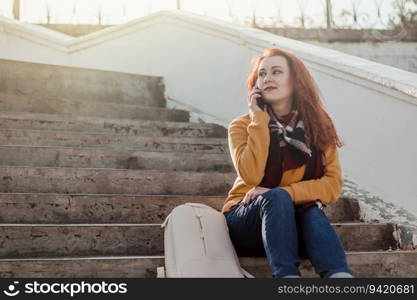 Young red-haired woman sitting on stairsand talking on phone. Lady in yellow sweater uses smartphone on sunny day. Lifestyle, copy space.. Young red-haired woman sitting on stairs and talking on phone. Lady in yellow sweater uses smartphone on sunny day. Lifestyle