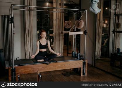 Young redhead fitness woman sitting with closed eyes in lotus position trying to meditate and relax after hard workout on pilates cadillac reformer in gym. Concept of healthy lifestyle and wellness. Young fit woman sitting in lotus position on trapeze table