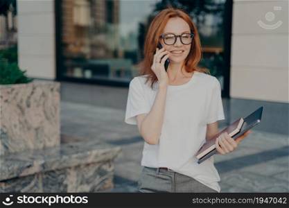 Young redhead smiling businesswoman talking on mobile phone outdoors, professional woman in casual clothes walking on city street and consulting client smartphone, holding modern laptop in hand. Young redhead smiling business woman in spectacles talking on mobile phone outdoors