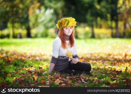 young redhead teenager woman in a wreath of maple leaves sitting on the grass