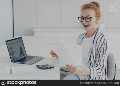 Young redhead woman feeling overjoyed while reading letter with bank notification or news about last mortgage payment, sitting at desk with opened laptop and calculator, female receiving money refund. Young redhead woman feeling overjoyed while reading letter with bank notification or news about last mortgage payment