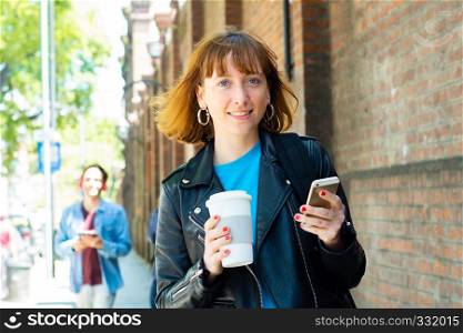 Young redhead woman holding coffee and reading message on his phone. Outdoors.