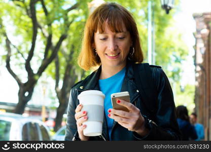 Young redhead woman holding coffee and reading message on his phone. Outdoors.