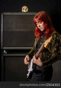 Young redhead woman playing electric guitar in front of large speaker cabinets.