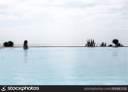 Young redhead woman swimming in an infinity pool in bikini near the ocean looking far away