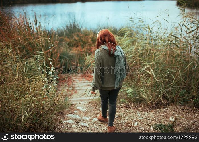 Young redhead woman walking to the lake shore with headphones wearing a jersey and black jeans