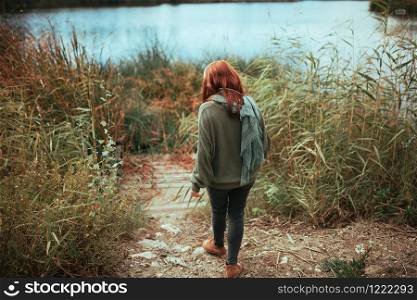 Young redhead woman walking to the lake shore with headphones wearing a jersey and black jeans