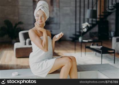 Young relaxed beautiful young woman with makeup has perfect clean skin, applies moisturizing cream on body lotion, poses wrapped in towel at living room, looks happily aside. Wellness concept
