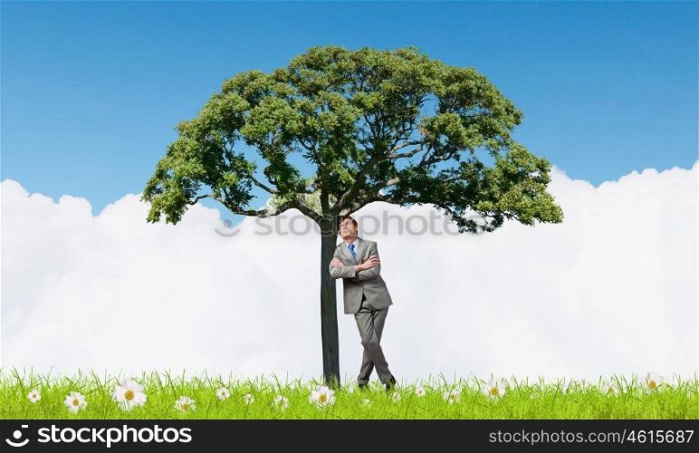 Young relaxed businessman leaning on green tree. Man and green tree