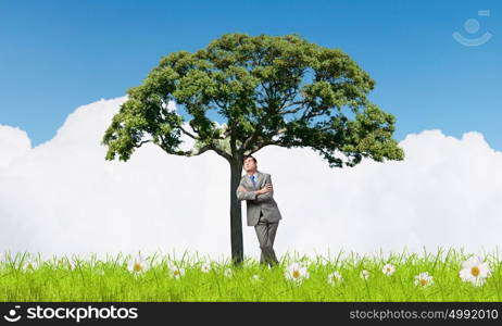 Young relaxed businessman leaning on green tree. Man and green tree