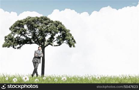 Young relaxed businessman leaning on green tree. Man and green tree