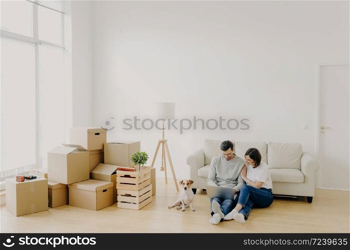 Young relaxed couple move to new home with their favourite pet, sit on floor, surf internet via laptop computer, have day of relocation, surrounded with cardboard boxes filled with personal belongins