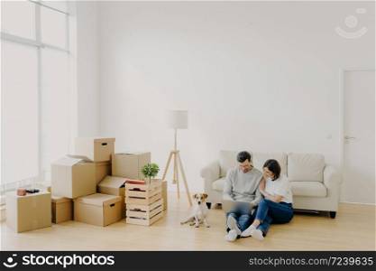 Young relaxed couple move to new home with their favourite pet, sit on floor, surf internet via laptop computer, have day of relocation, surrounded with cardboard boxes filled with personal belongins