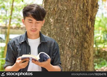 Young relaxed man reading book in nature, back on tree in garden