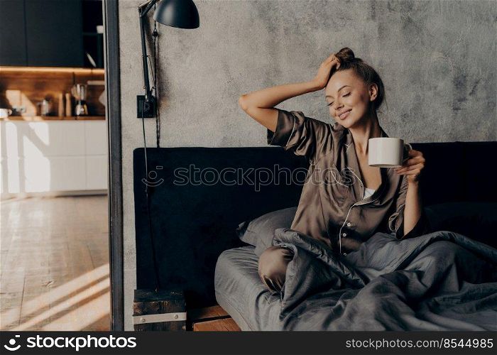 Young relaxed woman in stylish home suit waking up with cup of coffee in bed in cozy stylish bedroom at home, stretching from night sleep and enjoying morning leisure time and sunshine from window. Portrait of young relaxed woman in stylish home suit waking up with cup of coffee in bed