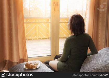 Young relaxed woman looking at the window. Young sexy woman sits relaxed on pillows with snacks near her and looks contemplative at the window, on a sunny day.