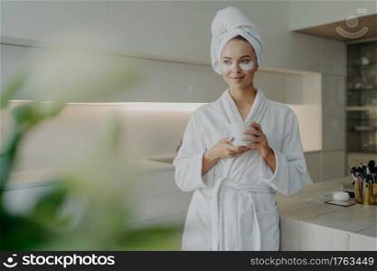 Young relaxed woman with cosmetic patches under eyes in bathrobe and hair wrapped in towel holding cup of tea and resting after spa treatments or taking bath while standing in modern kitchen at home. Beautiful woman in bathrobe and towel on head drinking tea while doing skin care procedures at home