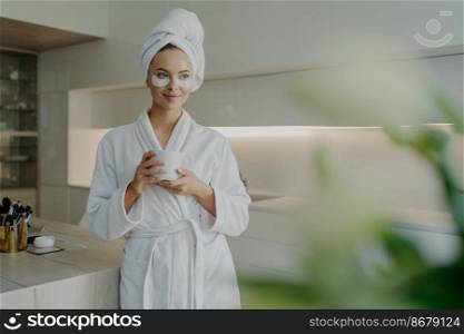 Young relaxed woman with cosmetic patches under eyes in bathrobe and hair wrapped in towel holding cup of tea and resting after spa treatments or taking bath while standing in modern kitchen at home. Beautiful woman in bathrobe and towel on head drinking tea while doing skin care procedures at home