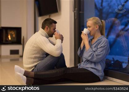Young romantic couple sitting on the floor in front of fireplace,talking and drinking tea on cold winter night at home