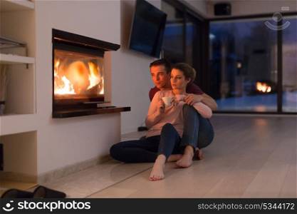 Young romantic couple sitting on the floor in front of fireplace,talking and drinking tea on cold winter night at home