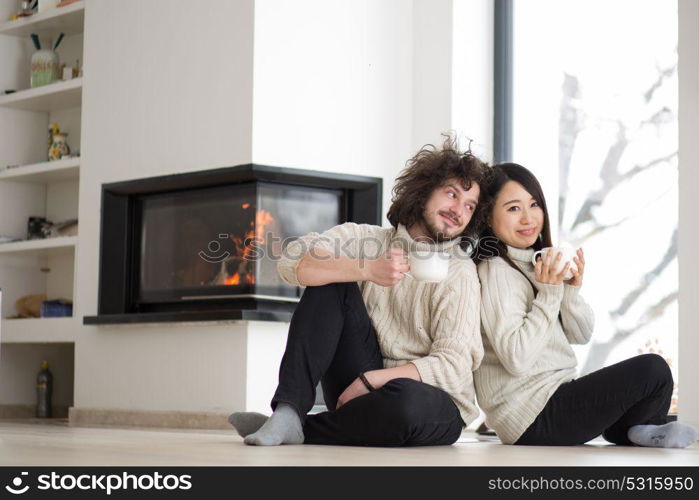 Young romantic multiethnic couple sitting on the floor in front of fireplace at home, talking and drinking coffee at cold winter day