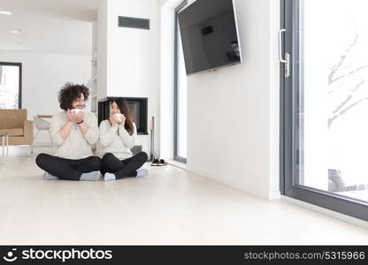 Young romantic multiethnic couple sitting on the floor in front of fireplace at home, talking and drinking coffee at cold winter day