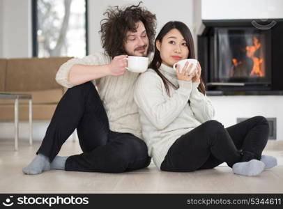 Young romantic multiethnic couple sitting on the floor in front of fireplace at home, talking and drinking coffee at cold winter day