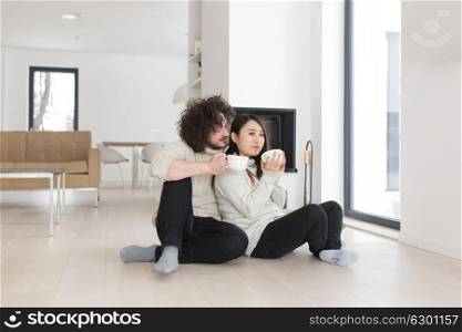 Young romantic multiethnic couple sitting on the floor in front of fireplace at home, talking and drinking coffee at cold winter day