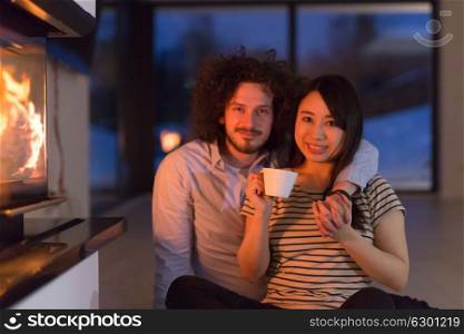 Young romantic multiethnic couple sitting on the floor in front of fireplace at home, talking and drinking tea on cold winter night