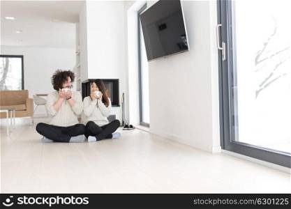 Young romantic multiethnic couple sitting on the floor in front of fireplace at home, talking and drinking coffee at cold winter day