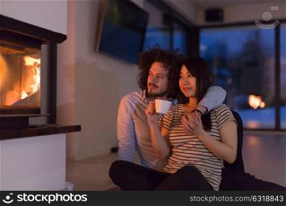 Young romantic multiethnic couple sitting on the floor in front of fireplace at home, talking and drinking tea on cold winter night